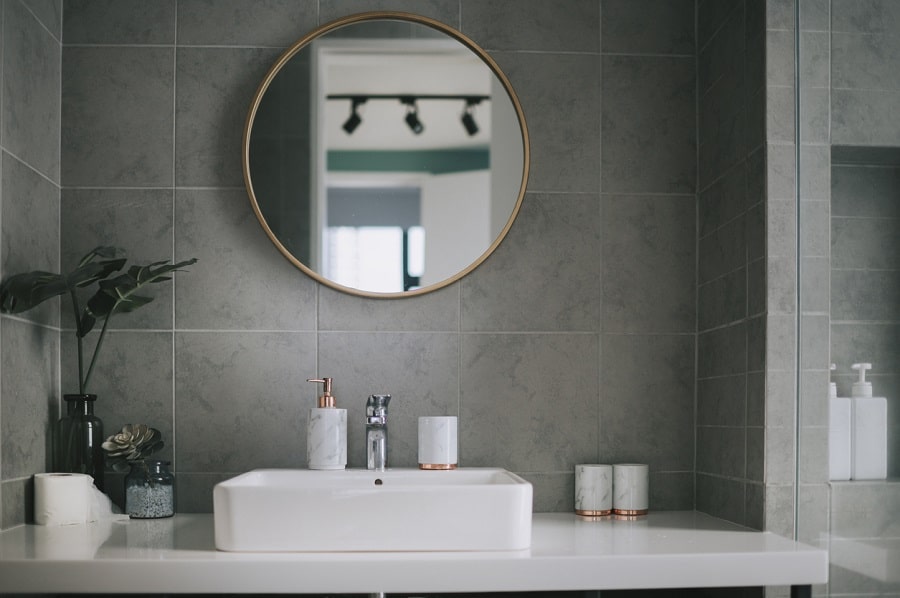 White countertop and rectangular sink below a round mirror on a gray tile wall